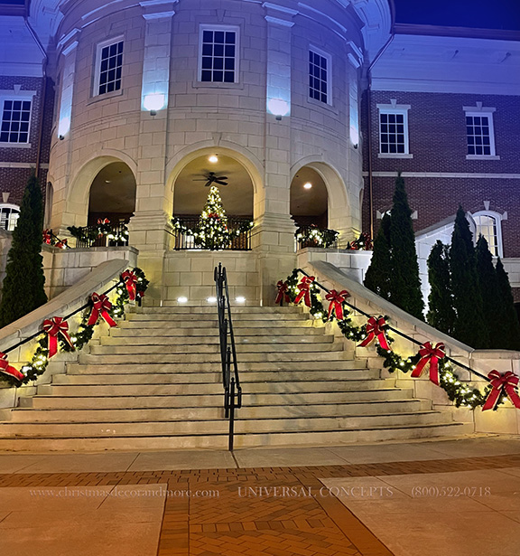 A view of a City Hall building with warm lighting and puffy red Christmas bows and draped garland.